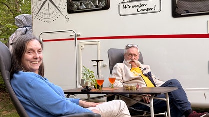 Das Bild zeigt die Erbslöhs vor ihrem Camper sitzen. Beide lächeln in die Kamera.