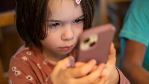 Ein Kind sitzt am Küchentisch und richtet seine ganze Aufmarksamkeit auf ein Smartphone.