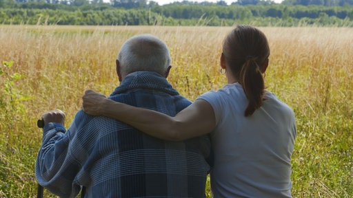 Ein älterer Herr sitzt mit seiner Tochter auf einer Bank vor einem großen Feld.