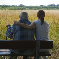 Ein älterer Herr sitzt mit seiner Tochter auf einer Bank vor einem großen Feld.