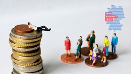 Ein Figürchen liegt auf einem Geldstapel, während andere Figuren auf kleineren Geldstapeln zu ihm hoch schauen