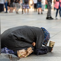 Eine Bettlerin kniet auf dem Boden, in den Händen hält sie einen Pappbecher, im Hintergrund bewegen sich Menschen auf der Domplatte.