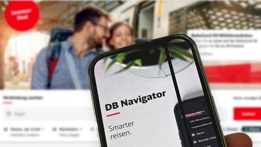 Eine Hand hält ein Handy mit der DB-App vor einem Desktop-Computer, auf dem die DB-Website geöffnet ist