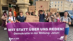 Schüler halten einen Banner mit der Aufschrift "Mit statt über uns reden! Demokratie in der Bildung: Jetzt!"