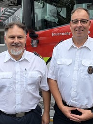 Zwei Männer in Feuerwehruniformen stehen vor einem Löschfahrzeug und lächeln in die Kamera