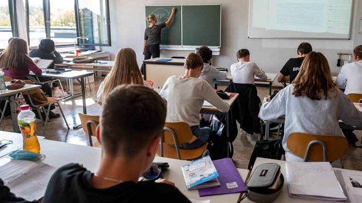 Symbolbild: Oberstufenschüler sitzen der Tafel zugewandt im Unterricht, die Lehrerin schreibt an die Tafel.