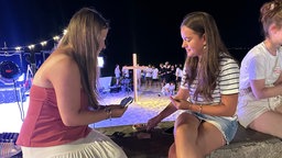 Zwei Frauen sitzen am Strand im Hintergund ist ein Holzkreuz zu sehen