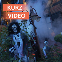 Ein Skelett steht als Halloween-Deko vor einem Haus. Im Hintergrund sieht man noch andere Dekorationen. 