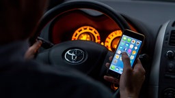 Ein Mann sitzt in einem Auto am Steuer und hält ein Smartphone in der Hand