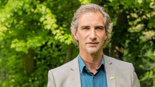 Porträtfoto eines Mannes mit grauen Haaren und gepflegtem Bart, der vor grünen Bäumen steht