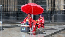 Zwei Karnevalisten sitzen bei regnerischem Wetter unter einem Sonnenschirm