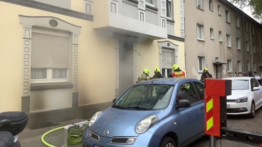 Schwarzer Rauch zieht aus einem Haus vor dem Einsatzkräfte der Feuerwehr stehen.