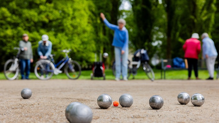Mehrere Menschen stehen in einem Park und spielen Boule, im Vordergrund liegen mehrere Kugeln