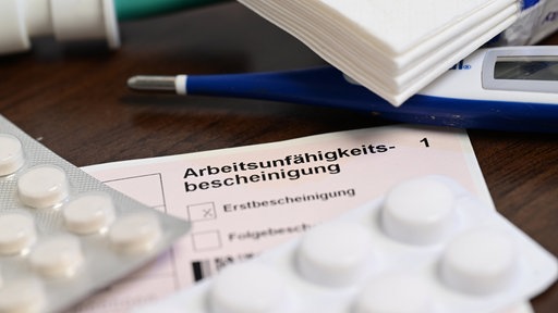 Auf einer Arbeitsunfähigkeitsbescheinigung liegen Tabletten und ein Fieberthermometer