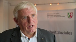  Karl-Josef Laumann spricht im WDR-Interview, im Hintergrund ist das Logo seines Ministeriums zu sehen