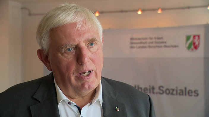  Karl-Josef Laumann spricht im WDR-Interview, im Hintergrund ist das Logo seines Ministeriums zu sehen
