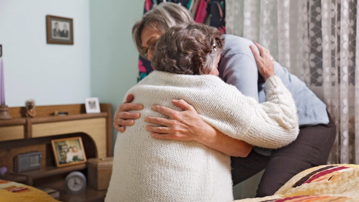 Ein Frau hat die Arme um eine ältere Frau geschlossen. Diese sitzt auf einem Bett und die jüngere Frau hilft ihr beim Aufstehen. Sie befinden sich in einem Schlafziimmer. Im Hintergrund stehen Bilderrahmen auf einer Holzkommode.