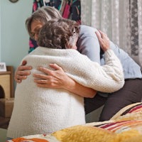 Ein Frau hat die Arme um eine ältere Frau geschlossen. Diese sitzt auf einem Bett und die jüngere Frau hilft ihr beim Aufstehen. Sie befinden sich in einem Schlafziimmer. Im Hintergrund stehen Bilderrahmen auf einer Holzkommode.