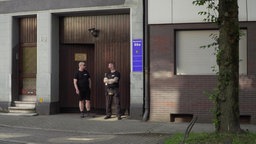 Ein braunes Holztor, davor zwei Männer, die die Tür bewachen. 