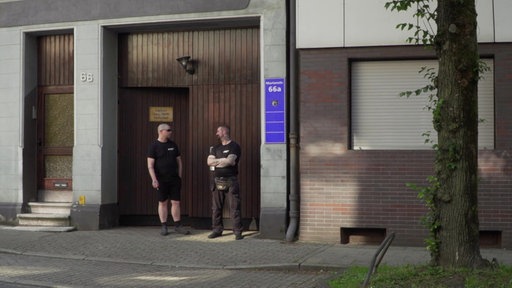 Ein braunes Holztor, davor zwei Männer, die die Tür bewachen. 