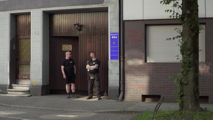 Es ist ein braunes Holztor zu sehen, davor stehen zwei Männer, die die Tür bewachen. 