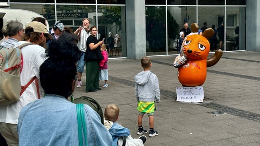 Menschen kommen vor der beschädigten Maus-Figur am Vierscheibenhaus in Köln zusammen 