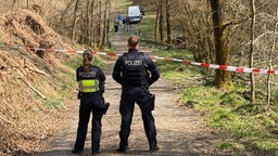 Auf einem Waldweg sind zwei Polizisten von hinten vor einem rot-weißen Flatterband zu sehen. Weit im Hintergrund arbeitet ein Team der Spurensicherung.