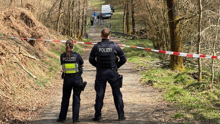 Auf einem Waldweg sind zwei Polizisten von hinten vor einem rot-weißen Flatterband zu sehen. Weit im Hintergrund arbeitet ein Team der Spurensicherung.