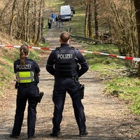 Auf einem Waldweg sind zwei Polizisten von hinten vor einem rot-weißen Flatterband zu sehen. Weit im Hintergrund arbeitet ein Team der Spurensicherung.
