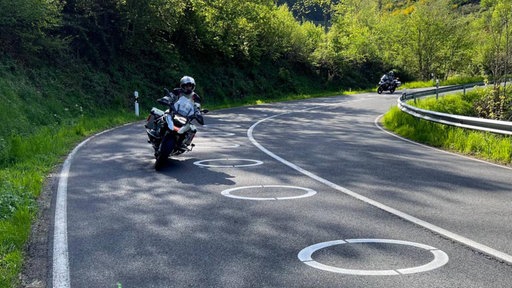 Blick auf die L218 im Kreis Düren, auf der Fahrbahn sind ellipsenförmige Markierungen, zwei Motorräder fahren in eine Kurve