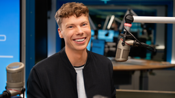 Das Bild zeigt den freien Journalisten Marek Walde, wie er vor einem Mikrofon in einem Radiostudio sitzt und in die Kamera lächelt. 