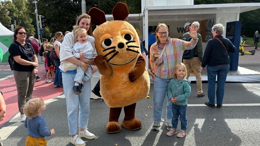 Die Maus steht in der Mitte und winkt, daneben stehen Eltern und Kinder und winken mit