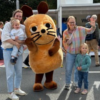 Die Maus steht in der Mitte und winkt, daneben stehen Eltern und Kinder und winken mit