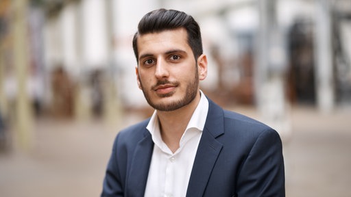 Ein junger Mann im Anzug, weißem Hemd, mit dunklen Haaren und Bart