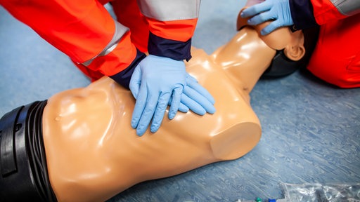 Zwei Hände mit blauen Gummi-Handschuhe pressen bei einer Herz-Druck-Massage im Rahmen eines erste Hilfe-Kurses auf den Brustkorb einer Reanimationspuppe