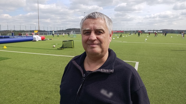 Ein älterer Mann steht lächelnd auf einem Fußballfeld