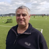 Ein älterer Mann steht lächelnd auf einem Fußballfeld