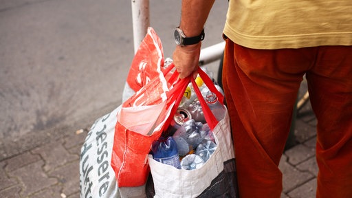 Ein Mann mit roter Hose und gelbem Shirt hält zwei Plastiktüten, die mit leeren Dosen und Plastikflaschen gefüllt sind.