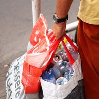 Ein Mann mit roter Hose und gelbem Shirt hält zwei Plastiktüten, die mit leeren Dosen und Plastikflaschen gefüllt sind.