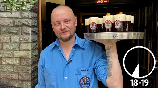 Köbes Patrick Lehnardt an seinem Arbeitsplatz. Er steht mit einem Tablett in der Hand vor der Tür des Hauses.
