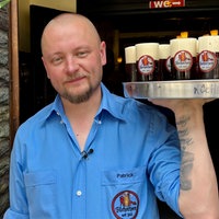Köbes Patrick Lehnardt an seinem Arbeitsplatz. Er steht mit einem Tablett in der Hand vor der Tür des Hauses.