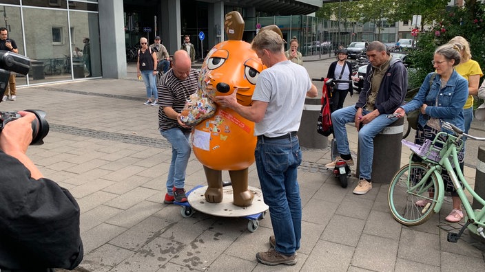 Die Maus wird auf einem Rollbrett von zwei Männern abtransportiert. Um sie herum stehen Menschen.