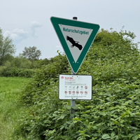 Ein Naturschutzgebietschild mit Verboten darunter steht auf einer grün bewachsenen Naturfläche. 