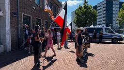 Eine kleine Gruppe an Menschen stehen vor einem Polizeiauto und schwingen eine schwarz, weiß, rot Flagge 