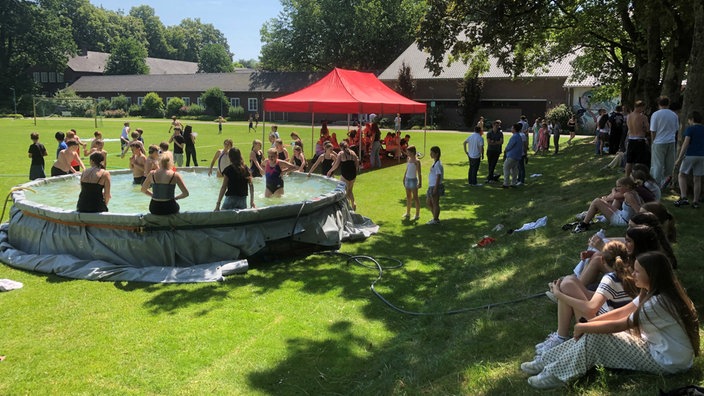 Versammlung einiger Schüler um einen runden Palettenpool auf der grünen Wiese des Schulhofs, bei Sonnenschein und klarem Himmel