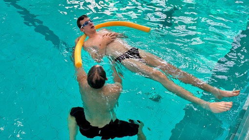 Ein Schwimmlehrer bringt einem Schüler mit einer orangenen Poolnudel schwimmen bei