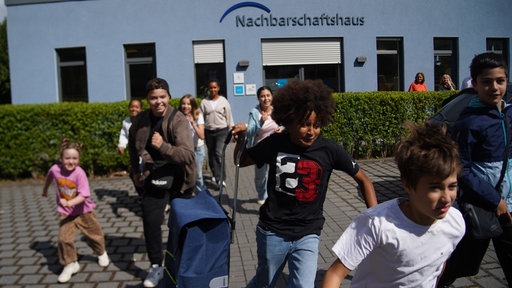 Die Kreativ Kids rennen mit ihrem Einkaufstrolley zur Kamera vor dem blauen Nachbarschaftshaus