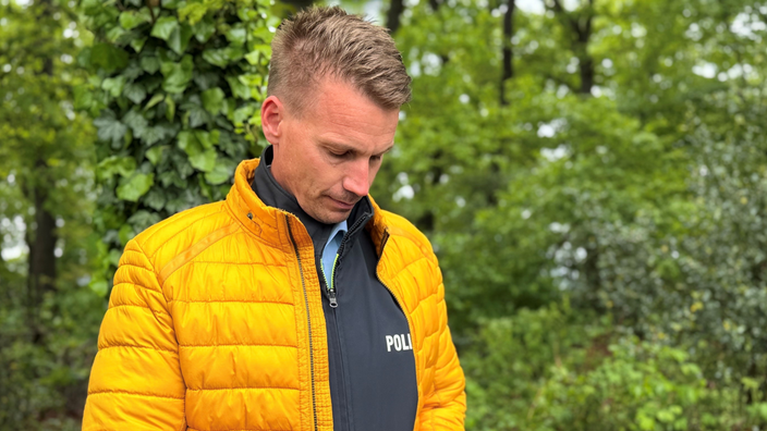 Der Polizist Christoph Neuhaus steht mit seinem Handy in der Hand da.