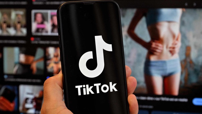 Tik Tok auf einem Smartphone vor einem Computerbildschirm, auf dem das Bild einer schlanken Frau zu sehen ist