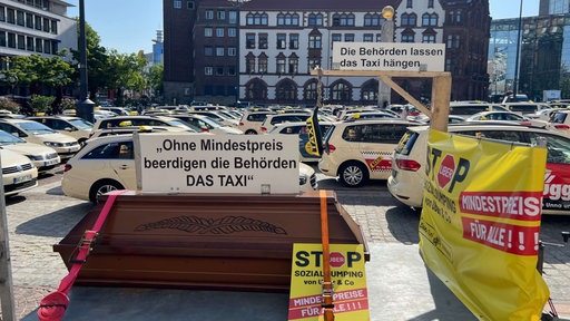 Auf dem „Parkplatz Friedensplatz“ vor dem Dortmunder Rathaus stehen viele Taxis. Im Vordergrund ist ein Sarg auf einem Anhänger zu sehen. Daruf steht ein Schild mit der Aufschrift: "Ohne Mndestpreis beerdigen die Behörden DAS TAXI"